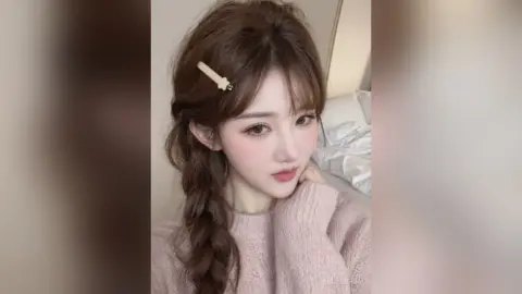 Young woman with braided hair and cream sweater posing in a cozy bedroom setting.