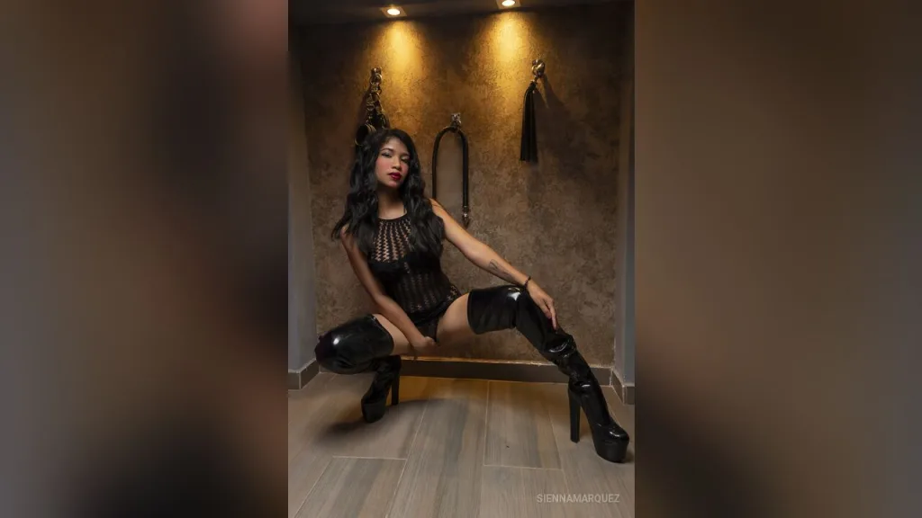 SiennaMarquez — SiennaMarquez poses in a black mesh bodysuit and tall black platform boots in a dimly lit room.