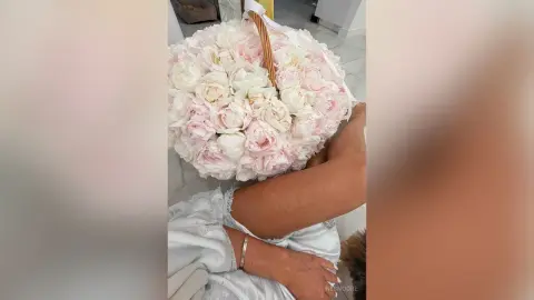 Woman in white lace robe holding a large basket of soft pink peonies on a marble floor.
