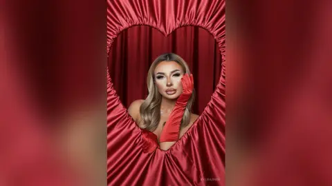 Glamorous woman in red evening gown and long gloves posing through a heart-shaped red curtain frame.
