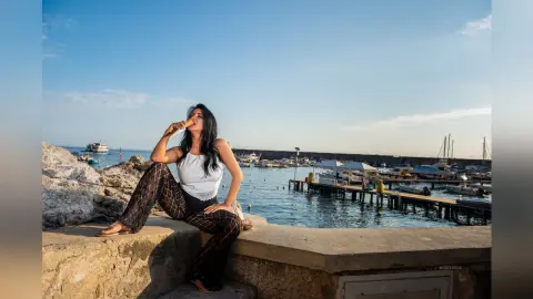 MissyJolie sits on a stone ledge by the ocean wearing a white lace top and black patterned leggings.