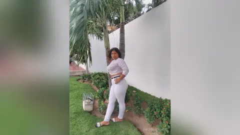 Stylish woman in a white striped crop top and white jeans posing in a sunny tropical garden setting.
