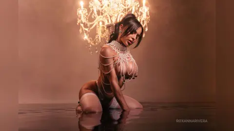 Glamorous woman in sparkling crystal body jewelry posing under a warm chandelier in a moody, elegant setting.