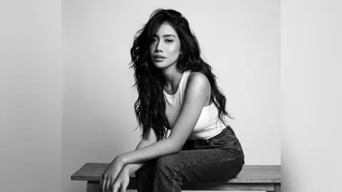 Elegant woman in a white tank top and jeans sitting against a minimalist background in a moody black and white portrait.