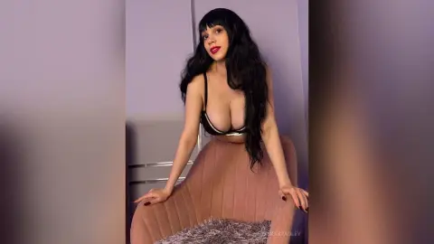 Smiling woman with long dark hair in black lingerie sitting on a pink velvet chair in a soft lit room.
