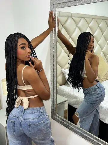 Young woman with long braids in a beige crop top and denim jeans posing in a bedroom mirror reflection.