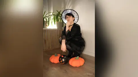Young woman in a witch costume and hat sitting by pumpkins in a cozy indoor setting for Halloween.