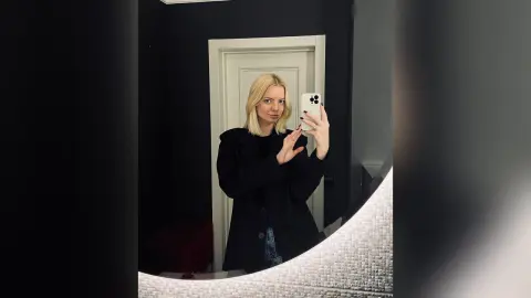 Blonde woman in a black blazer taking a moody mirror selfie in a modern dark room.