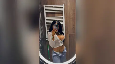 Young woman in a white crop top and denim jeans taking a mirror selfie in a modern bathroom.