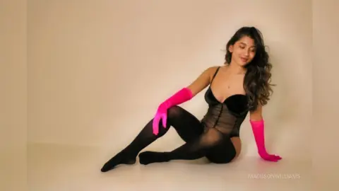 MadiisonWilliams poses in a black sheer bodysuit and long pink gloves, sitting on the floor against a plain white background.