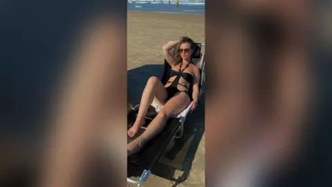 Relaxed woman in a black cutout swimsuit lounging on a beach chair during a sunny summer day at the seaside.