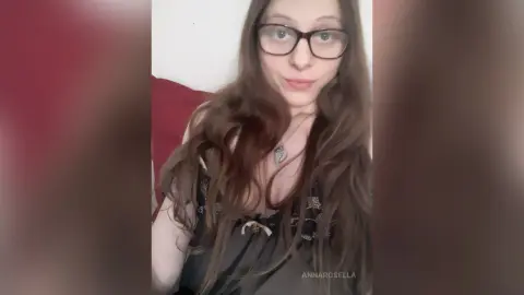 Smiling young woman with long wavy hair and glasses wearing a black patterned tank top in a cozy indoor setting.