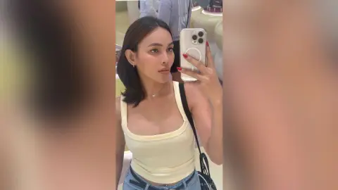 Young woman in a white tank top taking a mirror selfie in a bright, modern boutique setting.