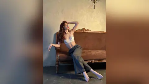 Elegant woman in white lace lingerie and dark trousers relaxing on a vintage brown sofa in a moody studio.