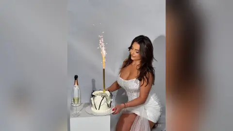 Woman in white sequin party dress celebrating with a birthday cake and sparkling candle in a bright studio.