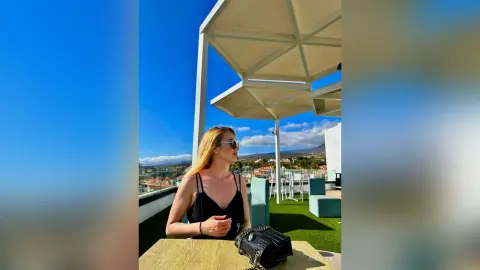 AmaFox — Blonde woman in a black tank top sitting at a rooftop cafe under a white canopy on a sunny day.