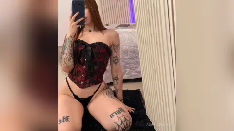 Woman in red lace corset and black lingerie posing for a mirror selfie in a modern bedroom.
