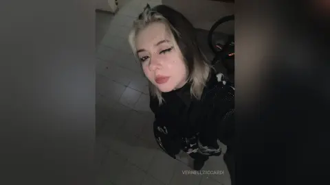 VernellZiccardi looks at the camera with blonde-tipped hair, wearing a black patterned top in a room with tiled flooring.