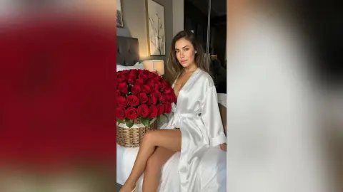 Smiling woman in a white satin robe sitting on a bed next to a large bouquet of red roses in a cozy bedroom.