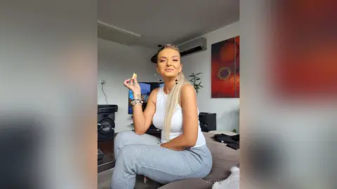 DelliaSmith wearing a white tank top and grey leggings, sitting on a couch while holding a small piece of food.