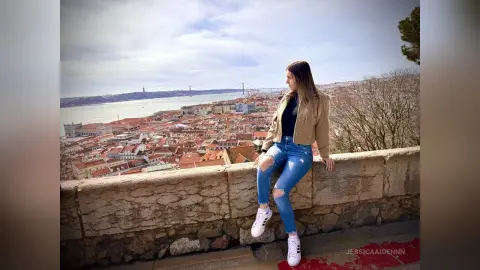 JessicaAidennn — Young woman in jeans and sneakers sitting on a stone wall overlooking the scenic Lisbon cityscape.