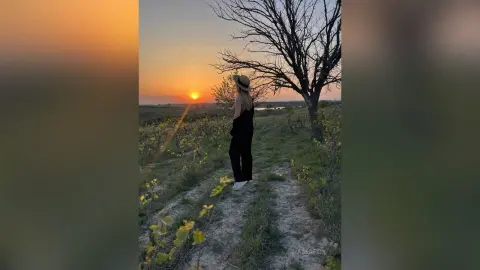 Woman in black outfit and sun hat gazing at a golden sunset over a rural vineyard landscape.
