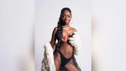 Smiling Black woman in black lingerie and a white faux fur coat posing against a plain studio background.