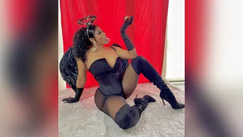 Woman in black angel costume with wings and halo posing playfully on a white rug against red curtains.