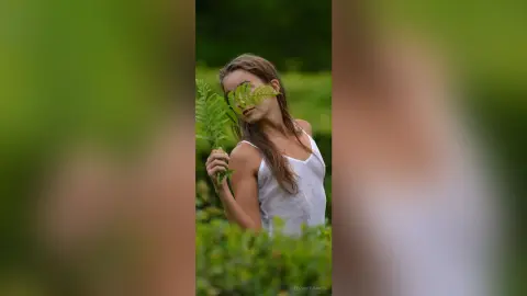 Dreamy portrait of a woman in a white tank top holding a green fern leaf in a lush summer garden.