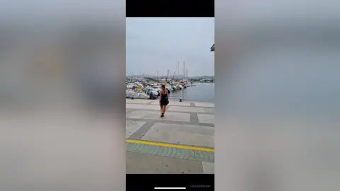 Woman in a black dress walking along a sunny marina pier near white sailboats and yachts.
