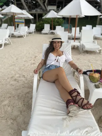 Relaxed woman in a white crop top and sun hat lounging on a beach chair under white umbrellas.
