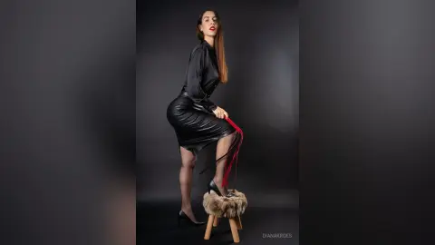 Elegant woman in a black satin blouse and leather skirt posing on a stool against a dark studio background.