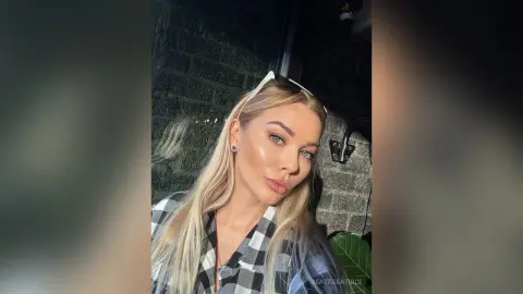 Blonde woman in a black and white checkered shirt posing outdoors at night with a glowing complexion.