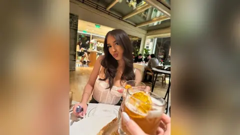 Young woman in a white lace top enjoying a refreshing cocktail at a bright, modern outdoor cafe.