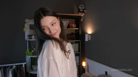 Smiling young woman in a white linen shirt relaxing in a cozy, dimly lit modern bedroom.