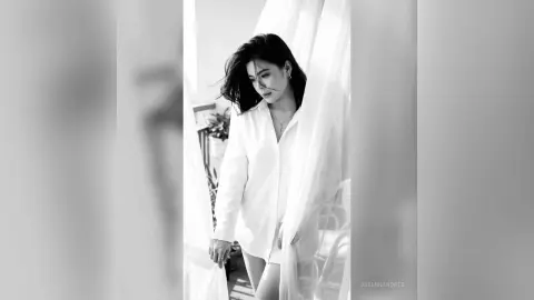 JasminAndres poses in a white button-down shirt and shorts by a bright window with sheer white curtains.
