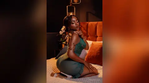 Elegant Black woman in a green satin dress posing on a bed in a moody, dimly lit bedroom setting.