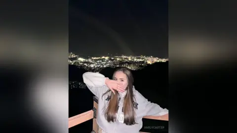 Young woman in a grey sweatshirt posing on a balcony overlooking a glowing city skyline at night.