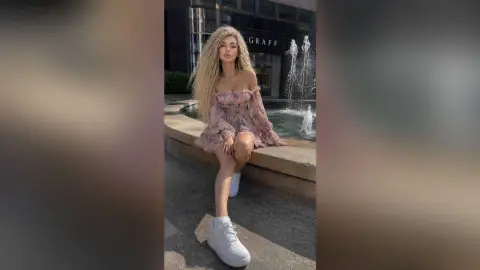 Blonde woman in a pink floral off-the-shoulder dress sitting by a city fountain in a stylish summer outfit.