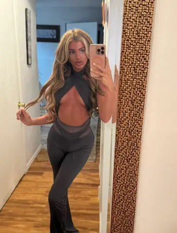 Blonde woman in a chic black cutout jumpsuit posing for a mirror selfie in a modern home hallway.