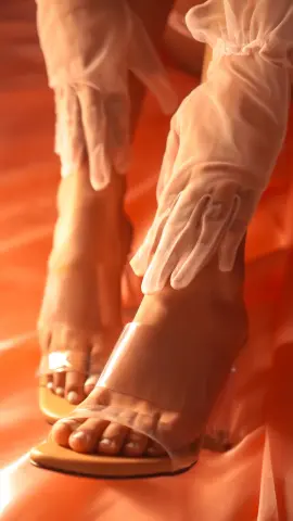 Elegant woman in sheer white gloves and clear high heel sandals posing on soft orange fabric.