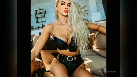 Blonde woman in black lingerie and high-waisted shorts sitting on a sofa in a bright, modern room.