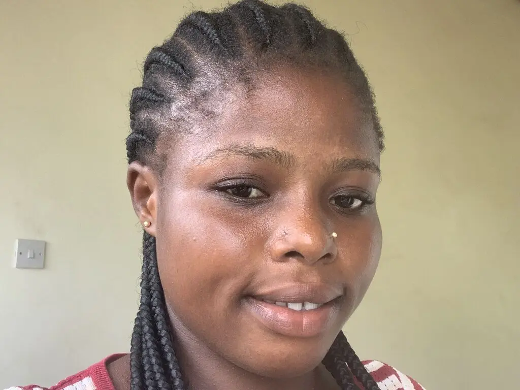 NancyEdward smiles close-up, showing her braided hair and nose piercing against a plain wall.