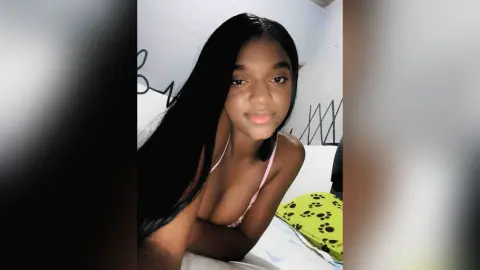 Young woman with long black hair wearing a pink bikini top in a bright bedroom setting.