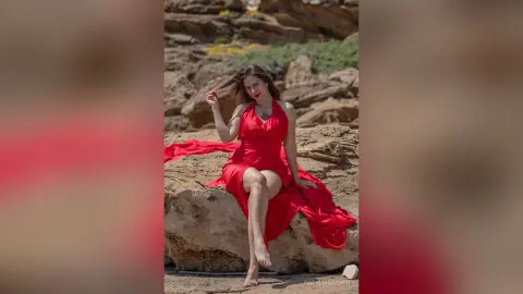 NataliaNacher poses outdoors in a long red dress, sitting on a rock with her long dark hair down.