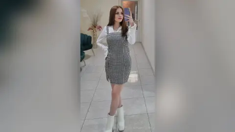 Young woman in a black and white gingham pinafore dress and white boots posing in a bright modern room.