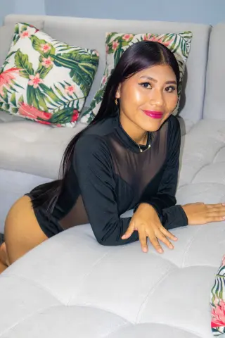Smiling young woman in a black long-sleeve sheer top relaxing on a white sofa with tropical floral pillows.
