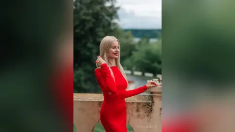 Smiling blonde woman in a long red dress posing on a stone balcony in a lush green garden setting.
