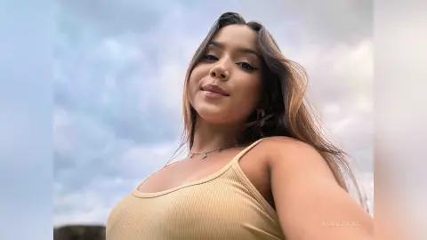 Young woman in a tan ribbed tank top smiling against a cloudy sky background.