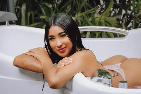 Smiling woman in white lace lingerie relaxing in a luxury outdoor white bathtub surrounded by green plants.